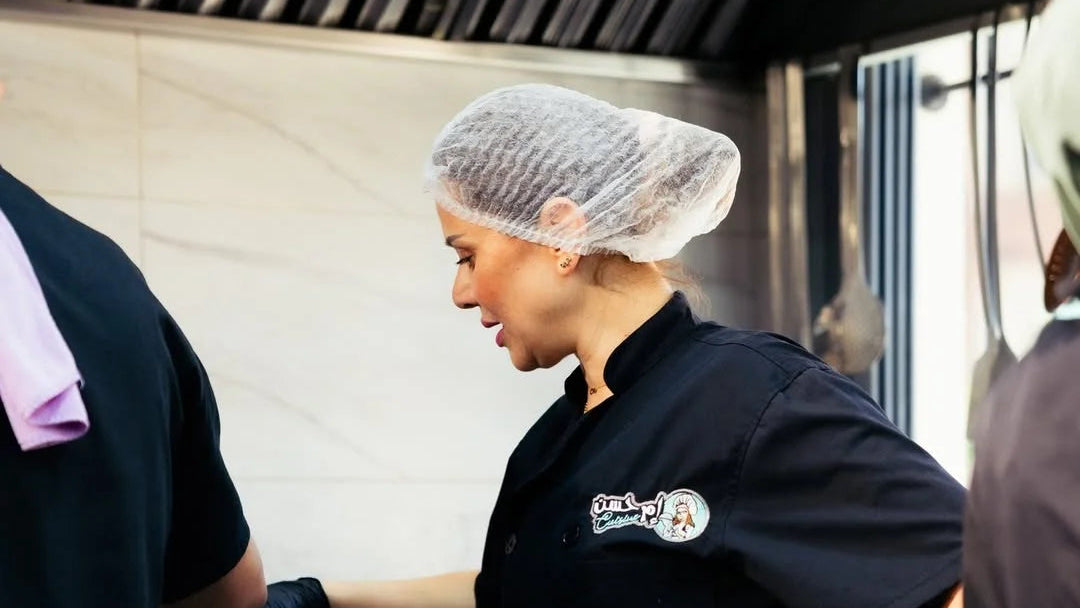 Person in a kitchen wearing a hairnet and black chef's coat with a logo.